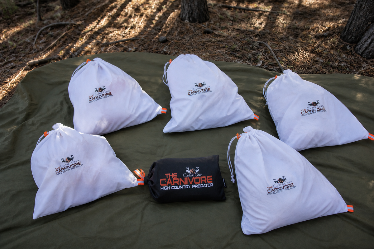 Five white Carnivore boned-out elk game bags and a black branded storage package arranged on a green tarp in shaded timber.