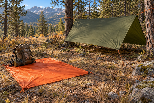 Compact orange tarp laid flat beside a backpack and a larger green tarp pitched between trees in Western mountain terrain.