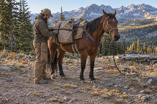 A packer checks a canvas side pannier on a horse along a rocky mountain trail.