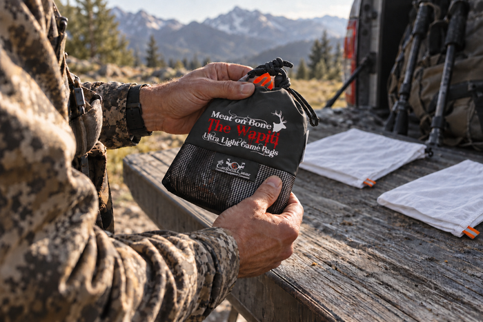 A hunter holds a black Wapiti game bag set pouch beside two folded white game bags on a truck tailgate at a mountain trailhead.