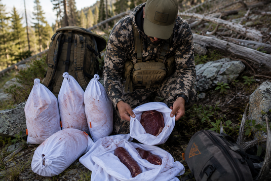 Hunter organizing elk backstraps and tenderloins in breathable white synthetic game bags with narrow orange side tabs during a mountain elk packout