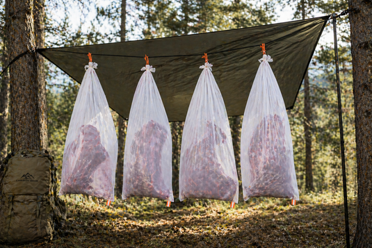 Elk quarters hanging in breathable white game bags with narrow orange reflective tabs under a tarp in shaded forest during early season hunt