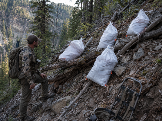 Hunter choosing an elk meat staging spot in shaded timber on a steep rocky slope during a multi-trip packout